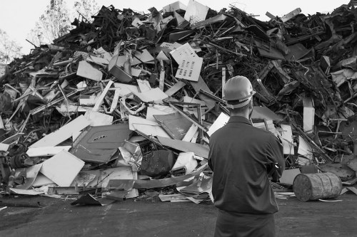 A man wearing a safety helmet and dark work clothing stands in the foreground, facing a large pile of mixed construction and household waste. The heap comprises various materials including broken furniture, plastic containers, cardboard, wood, metal scraps, and other debris, some of which are stacked haphazardly. The waste appears to be on an outdoor paved surface, possibly part of a rubbish disposal or clearance site in Southgate or nearby. The background shows a few trees and a partly visible fence, indicating an open storage area. The scene is captured in black and white, emphasizing the textures and disorder of the waste materials, which Office Clearance Southgate may assist with removing from small businesses or residential properties as part of their rubbish clearance services.