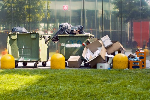 In the foreground, three large green wheelie bins with black lids are positioned on a paved surface, partially covered with black plastic trash bags. To the right of the bins, there are discarded cardboard boxes, some flattened and others partially assembled, with visible tape and printed labels. The boxes vary in size and are scattered among orange and blue plastic crates, which contain miscellaneous waste items. Yellow bollards are positioned in front of the waste containers, separating them from a well-maintained green grassy area that lines the paved surface. Behind the waste, a modern glass building with reflective windows reflects trees and the surrounding environment, indicating an urban location. The overall scene suggests a cleared area for rubbish disposal, consistent with professional waste management and rubbish removal services such as those offered by Office Clearance Southgate, situated near Southgate or associated postcode areas.