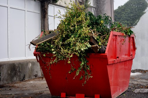 A large red plastic skip filled with green garden waste, including leafy branches and foliage, positioned outdoors on a paved surface next to a white fence and concrete wall. The skip's sides are slightly inclined, showing the contents piled high, with some branches spilling over the edge. In the background, there is a grey wall with climbing plants and a small section of greenery visible beyond the fence. The lighting suggests natural daylight, highlighting the textures of the leafy waste and the smooth surface of the skip, which is likely used for rubbish removal services in Southgate. This scene illustrates a typical outdoor clearance process managed by Office Clearance Southgate, specializing in domestic and commercial rubbish disposal projects in the local area.