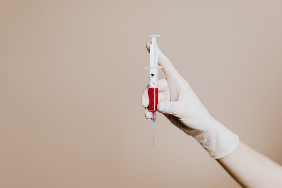 A close-up image of medical waste items arranged on a light grey surface, including two disposable syringes with clear barrels and black measurement markings, a small amber glass bottle with a white s