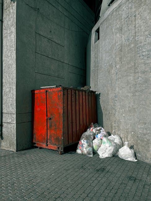 A red metal waste container with a weathered finish, positioned against a dark teal wall and a light grey concrete wall on a paved surface. The container is rectangular with vertical ridges along its 
