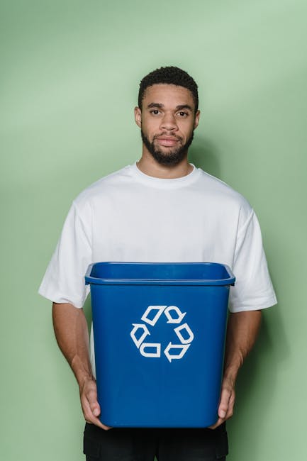 A young man with short, curly dark hair and a beard, wearing a plain white T-shirt, is standing against a solid light green background, holding a blue recycling bin with a white recycling symbol on th