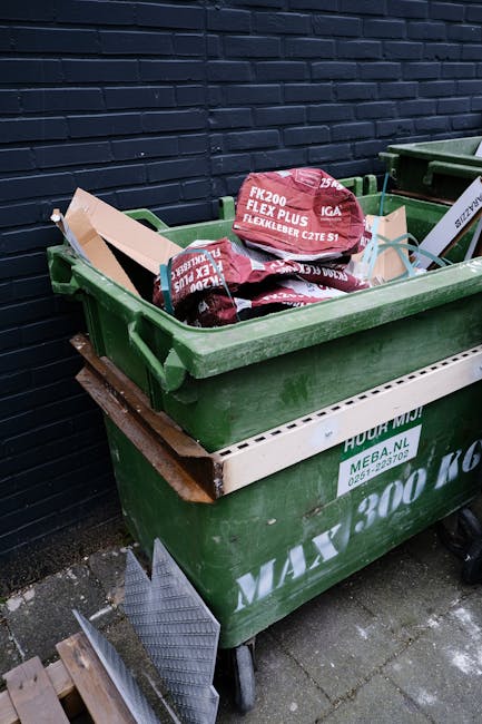 A row of multiple wheeled bins placed outdoors along a pavement, with green, blue, and red hoppers, each with closed lids and textured surfaces, positioned against a brick wall in a residential or urb