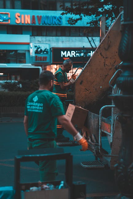 A sanitation worker dressed in bright yellow protective overalls and a matching helmet stands at the rear of a large industrial rubbish collection truck during daytime. The back of the truck is open, 